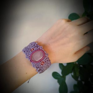Puede incluir: Pulsera de macramé artesanal en tonos lavanda y rosa. Presenta una piedra central ovalada de cuarzo rosa, rodeada de piedras rosas más pequeñas y un intrincado trabajo de nudos. La pulsera está diseñada para una muñeca.