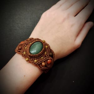 Puede incluir: Una pulsera de macramé marrón con una gran piedra verde ovalada en el centro. La pulsera está adornada con piedras más pequeñas, redondas y ovaladas, en tonos marrones y rojos. La pulsera está en una muñeca sobre un fondo oscuro.