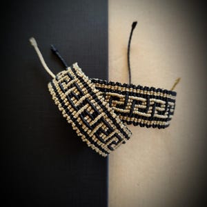 Pode incluir: Duas pulseiras de macramé com um padrão geométrico em preto e bege. As pulseiras são feitas de cordão tecido e têm um design clássico. O fundo é dividido entre preto e bege.