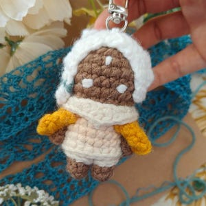 May include: Crocheted keychain featuring a small figure with a brown head, white hat, and cream-colored body. The figure has yellow arms and small brown feet. It is attached to a silver-colored metal clasp.