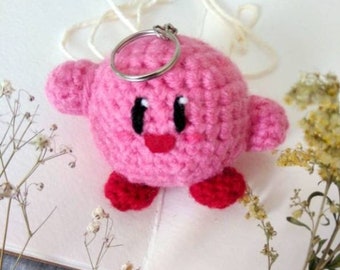 May include: Pink crocheted keychain shaped like a cartoon character with a key ring attached. The character has a round body, red feet, and a smiling face with black eyes.
