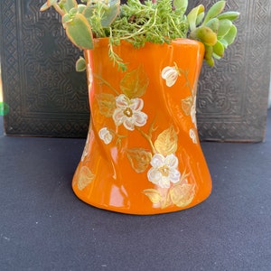 May include: A small orange ceramic planter with a white floral design and gold accents. The planter is filled with green succulents.