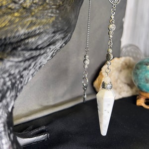 May include: A white and gray crystal pendulum with a silver chain and a silver charm hanging from a black raven figurine.