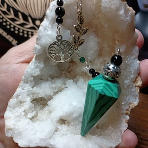 May include: A green malachite pendulum with a silver chain and a tree of life charm. The pendulum is hanging from a white crystal geode.