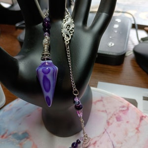 May include: A purple and white agate pendulum with a silver chain and beads. The pendulum is hanging from a silver ornate charm.