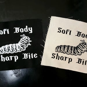 May include: Two square patches, one black and one off-white, each featuring a white illustration of a caterpillar and the text "Soft Body Sharp Bite" in a gothic font. The patches are likely for clothing or accessories.