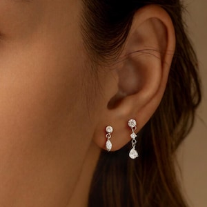 Könnte beinhalten: Nahaufnahme von zwei Paar silbernen Ohrringen mit klaren, funkelnden Edelsteinen. Ein Paar hat einen tropfenförmigen Stein, das andere eine Ansammlung und einen hängenden Tropfen. Die Ohrringe sind vor einem warmen, neutralen Hintergrund.