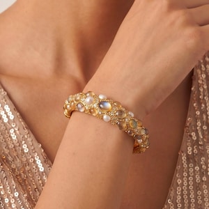 Könnte beinhalten: Ein goldfarbenes Armband mit einer strukturierten Metalloberfläche, die mit zahlreichen runden, schillernden, mondsteinartigen Cabochons und kleinen weißen Perlen besetzt ist, welche in einem dichten Muster auf dem Band angeordnet sind.