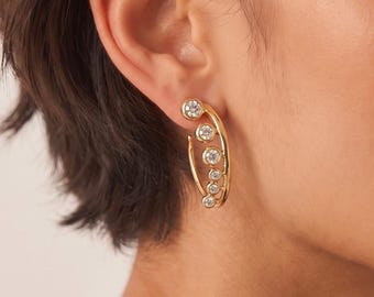 May include: A gold ear climber earring featuring a curved metal frame set with five round, clear, bezel-set stones that graduate in size from the top to the bottom, following the natural contour of the earlobe and cartilage.