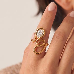 Könnte beinhalten: Ein goldfarbener Ring mit einem fließenden, wellenförmigen Drahtdesign, das sich um den Finger legt. Der Ring ist mit einem einzelnen, klaren, birnenförmigen Edelstein akzentuiert, der in der oberen Schlaufe der Metallstruktur eingefasst ist.