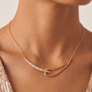 Könnte beinhalten: Eine goldene Halskette mit einer feinen Kette und einem zentralen Anhänger aus zwei ineinandergreifenden, geschwungenen Elementen. Ein Element ist ein glatter, polierter Goldbogen, während das andere mit kleinen, klaren, funkelnden Steinen besetzt ist.