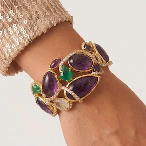 Könnte beinhalten: Ein Goldarmreif mit mehreren ovalen violetten Amethysten und grünen Smaragden, die in einen mit kleinen weißen Pavé-Diamanten besetzten Rahmen gefasst sind. Das Armband hat eine offene, organische Struktur, die die verschiedenen Steine verbindet.