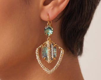 May include: A gold-tone drop earring featuring a small, iridescent abalone shell stone at the top, connected to a larger, geometric, open-frame pendant. The frame is encrusted with small clear crystals and holds a central, faceted abalone shell.