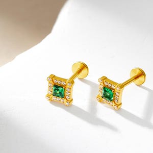 May include: A pair of gold stud earrings. Each earring has a square, emerald-green gemstone at the centre, encircled by smaller, clear gemstones. The earrings are presented on a white, textured surface.