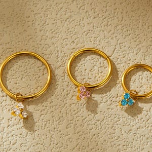 Könnte beinhalten: Vier goldfarbene Creolen mit kleinen, farbenfrohen Blumenanhängern. Die Anhänger haben weiße, rosa und blaue Edelsteine. Die Ohrringe werden auf einer strukturierten, hellen Oberfläche präsentiert.
