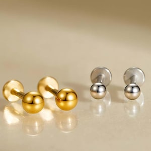 Könnte beinhalten: Eine Sammlung goldener und silberner Kugelstecker-Ohrringe. Die goldenen Ohrringe haben eine helle, polierte Oberfläche, während die silbernen Ohrringe ein mattes Aussehen haben. Die Ohrringe werden auf einer hellen Oberfläche mit weicher Beleuchtung präsentiert.