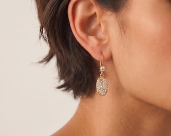 May include: A gold drop earring featuring a small round clear stone at the top and a larger oval-shaped pendant below. The oval pendant is encrusted with numerous tiny, sparkling clear stones set closely together in a textured gold frame.