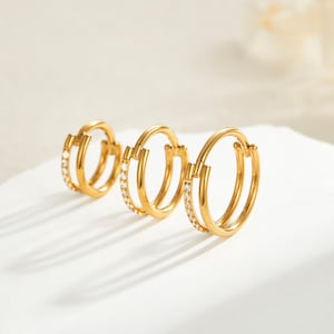 Könnte beinhalten: Drei goldfarbene Creolen-Ohrringe mit Doppelreifen-Design. Ein Reifen ist mit kleinen, klaren Edelsteinen besetzt. Die Ohrringe werden auf einer weißen Oberfläche präsentiert und werfen Schatten. Das Schmuckstück wird in einem Studio-Setting präsentiert.