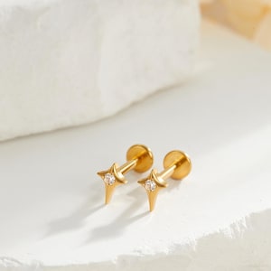 May include: A pair of gold-colored star-shaped earrings with a small, clear gemstone in the center of each star. The earrings have a post and flat back design. The earrings are displayed on a white surface.