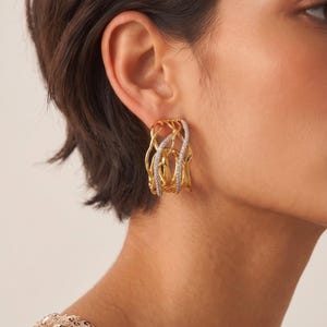 Könnte beinhalten: Ein goldener Creolen-Ohrring mit einem komplizierten, durchbrochenen Design aus ineinander verschlungenen, wellenförmigen Metallsträngen. Einer der Stränge ist mit kleinen, klaren, funkelnden Steinen besetzt, die einen Kontrast bilden.