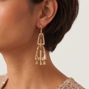 May include: A gold-toned drop earring featuring two interconnected geometric shapes with a hammered texture. The upper trapezoidal frame and the lower wider frame are both accented with small, clear pavé-set stones along their central vertical bars.