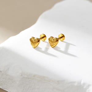 Könnte beinhalten: Zwei goldfarbene herzförmige Ohrstecker. Auf jedem Ohrring steht der Schriftzug "KISS ME". Die Ohrringe liegen auf einer weißen Oberfläche.