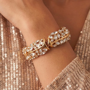 May include: A gold-toned hinged bangle bracelet featuring a repeating leaf-like pattern encrusted with clear, marquise-cut crystals. The metal framework is polished and holds the stones in a structured, symmetrical arrangement around the wrist.