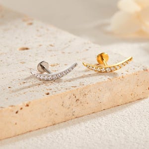 Op de afbeelding: Twee gebogen oorstekers. Eén is zilverkleurig en bezet met heldere kristallen, de andere is goudkleurig met heldere kristallen. De oorbellen zijn halvemaanvormig en hebben een pin en een sluiting voor doorboorde oren. Ze worden gepresenteerd op een getextureerd, licht oppervlak.