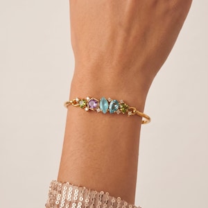 May include: A gold-toned bracelet featuring a central arrangement of multi-coloured gemstones, including a purple hexagon, a light blue marquise, and a teal pear-shaped stone, accented by small green stones and tiny clear crystals along the band.