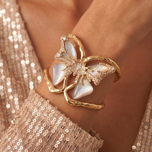 May include: A gold cuff bracelet featuring a large butterfly centrepiece with iridescent white shell wings and clear crystal accents. The metal band has a textured, organic finish that curves around the wrist to support the butterfly design.