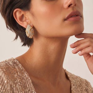 Könnte beinhalten: Ein ovaler Ohrring mit einem goldfarbenen Metallrahmen, der einen gewellten, erhabenen Rand aufweist und dessen Oberfläche dicht mit zahlreichen kleinen, klaren, funkelnden Kristallen besetzt ist, die das Licht reflektieren.