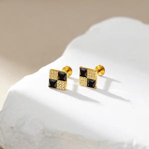 Könnte beinhalten: Ein Paar goldfarbene quadratische Ohrstecker. Jeder Ohrstecker hat ein Schachbrettmuster mit schwarzen und klaren quadratisch geschliffenen Steinen. Die Ohrringe werden auf einer weißen Oberfläche präsentiert.