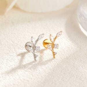 May include: Two leaf-shaped stud earrings, one silver and one gold, each adorned with clear, sparkling crystals. The earrings feature a delicate branch design with multiple crystal accents, perfect for adding a touch of elegance.