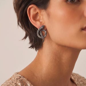 May include: A dark metal snake-shaped earring featuring a coiled design encrusted with numerous small, clear, sparkling stones. The earring is positioned on the earlobe, showcasing a detailed, textured surface with a metallic finish and shimmering accents.