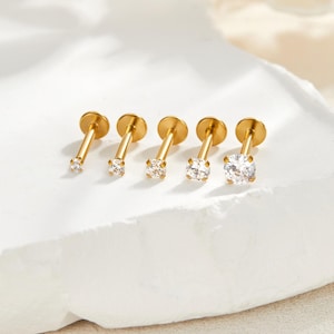 Könnte beinhalten: Fünf goldfarbene Lippenpiercing-Stecker mit klaren, runden Zirkonia-Steinen. Die Stecker sind in einer Reihe angeordnet, wobei die Steine von links nach rechts an Größe zunehmen. Die flache Rückseite jedes Steckers ist sichtbar.