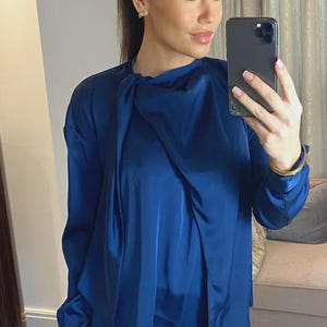 May include: A long-sleeved, royal blue blouse with a draped neckline. The blouse is made of a shiny, silky material and is paired with black leather-look trousers. The model is holding a black smartphone.