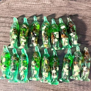 May include: A group of translucent green resin Virgin Mary figurines. Each figure contains a mix of natural elements, including white and brown inclusions. The figurines are displayed on a wooden surface, highlighting their detailed forms and vibrant colour.