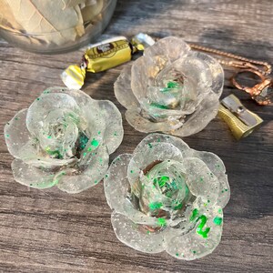 May include: Three clear resin rose-shaped ornaments with green glitter accents. The roses are arranged on a wooden surface.