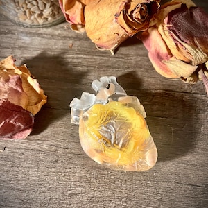 May include: A clear glass heart-shaped ornament with a yellow-gold interior. The ornament is sitting on a wooden surface with dried roses.