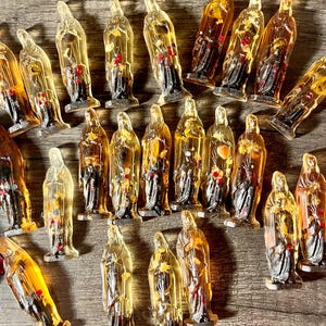 May include: A collection of small, translucent amber-coloured Virgin Mary figurines. The figures have a detailed design, with a dark base and red accents. The figurines are arranged on a wooden surface.