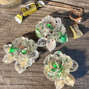 May include: Three clear resin rose flower decorations with green and gold accents. The flowers are on a wooden surface, along with a ring, a pencil sharpener, and a wrapped candy.
