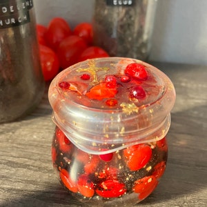 May include: A clear glass jar filled with red and black beads, gold flakes, and a small gold cross. The jar is sitting on a wooden surface.