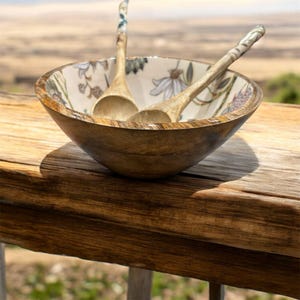 May include: A wooden bowl with a floral design and two wooden spoons inside. The bowl is sitting on a wooden surface.
