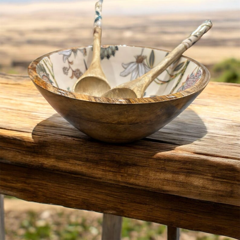 May include: A wooden bowl with a floral design and two wooden spoons inside. The bowl is sitting on a wooden surface.