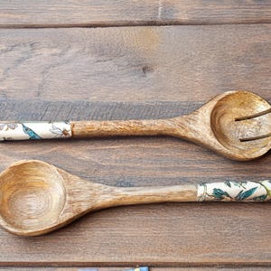 May include: Two wooden spoons with floral patterned handles. The spoon on the left is a serving spoon, while the spoon on the right is a slotted spoon.