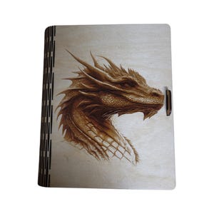 May include: A wooden box with a detailed dragon head design. The dragon is rendered in shades of brown, with intricate scales and sharp features. The box has a hinged side and a small clasp.