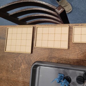 May include: Three small, light brown wooden game boards with grid patterns are arranged on a dark wooden surface. A metal tray with miniature figures is in the foreground. A dark brown metal chair is in the background.