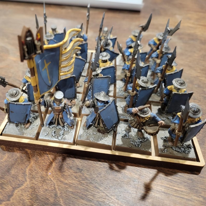 May include: Miniature figurines of a medieval army, featuring soldiers in blue and yellow uniforms, holding spears and shields. A banner with a cross is held high. The figures are arranged on individual bases within a wooden tray.