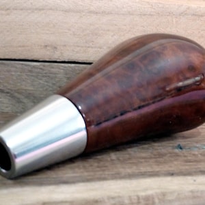 Może przedstawiać: Drewniany gałka zmiany biegów w kolorze brązowym z podstawą ze srebrnego metalu. Gałka ma unikalny wzór usłojenia drewna.