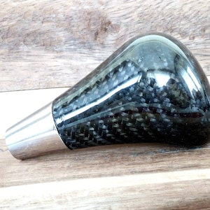 May include: A black carbon fibre gear shift knob with a silver metal base.